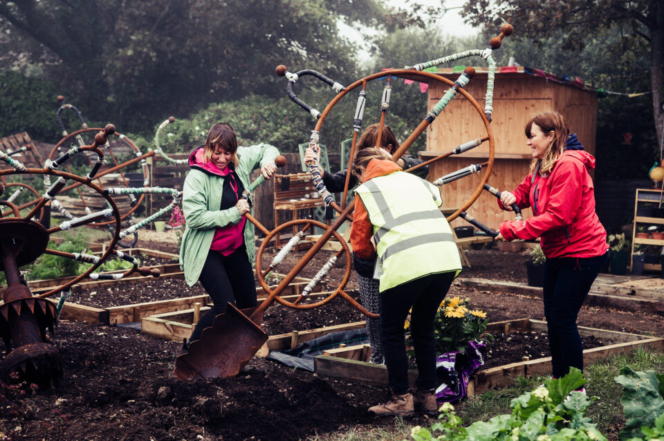 'Our Common Tools' by Katie Surridge b-side festival 2021. The photo depicts 4 people in an allotment carrying a very large tool like a shovel 