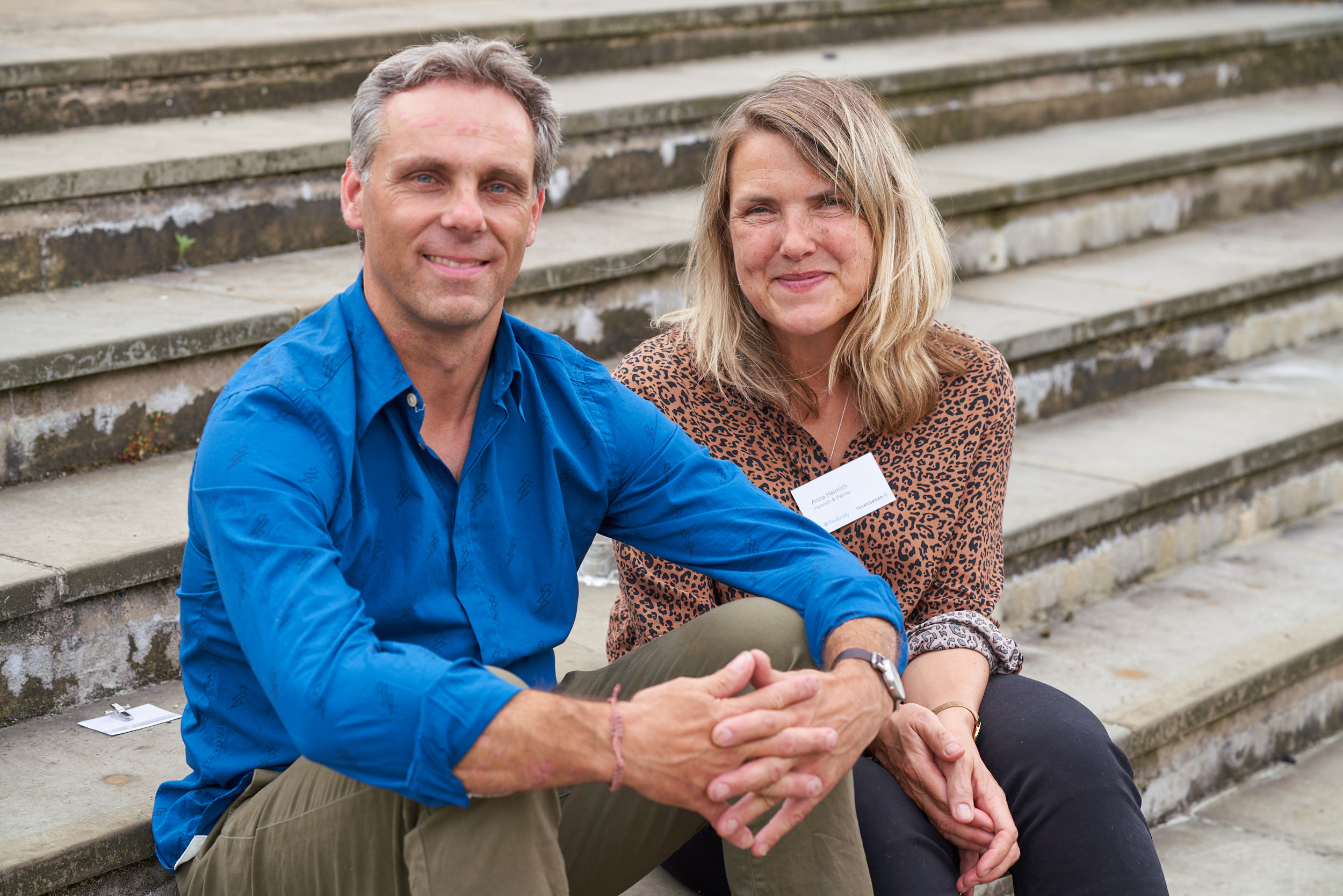 A photo of two people one male and one female. Sitting next to each other on some steps, they are smiling and looking towards the camera. The man is wearing a blue shirt, he has short light coloured hair and the woman is wearing a brown patterned shirts she has shoulder length blonde hair.